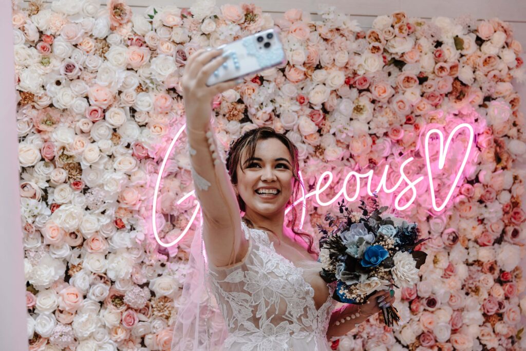 Bride taking a selfie in front of our pink and white flower wall.