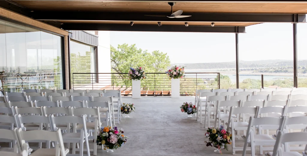 Outdoor wedding ceremony setup with rows of white chairs, colorful floral arrangements, and a scenic lake view in the background