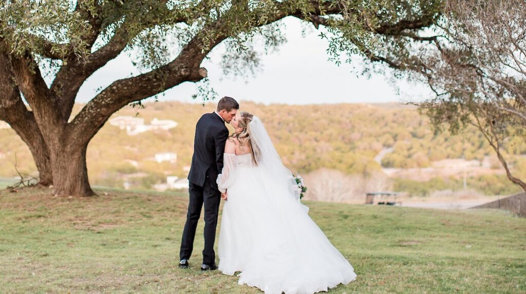 Romantic outdoor wedding portrait of the bride and groom kissing beneath a large, sprawling oak tree on a grassy hill overlooking a valley. 